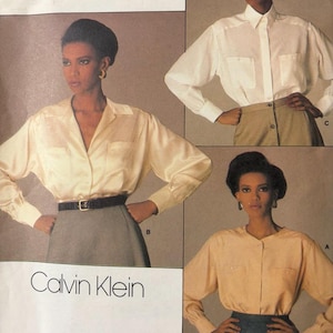 May include: A Vogue Patterns American Designer sewing pattern envelope for Calvin Klein, number 1455. The cover displays three variations of long-sleeved button-down shirts in white, cream, and peach, paired with skirts, featuring collars and chest pockets.