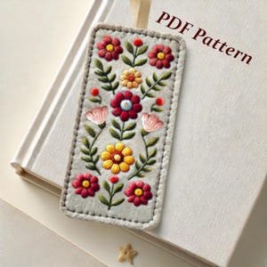 May include: A gray felt bookmark with an embroidered floral design. The flowers are red, yellow, and pink with green leaves. The bookmark has a white border and the text "PDF Pattern" is printed on the background.