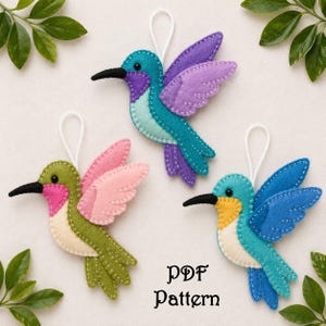 Peut inclure: Trois ornements en forme de colibris en feutrine, dans des tons de vert, rose, bleu et violet, chacun avec un bec noir et une ficelle blanche pour suspendre. Le texte "PDF Pattern" est en dessous.