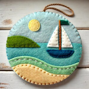 Könnte beinhalten: Ein Filz-Anhänger, der ein Segelboot auf einem blauen Meer mit einer grünen Insel und einer gelben Sonne zeigt. Der Anhänger ist rund und hat einen braunen Bindfaden zum Aufhängen.