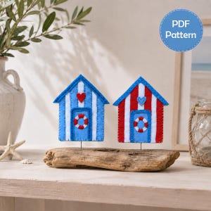 May include: Two felt beach hut ornaments mounted on a piece of driftwood. One hut features blue and white stripes with a red heart and life ring, while the other has red and white stripes with a blue heart and life ring. Text reads PDF Pattern.