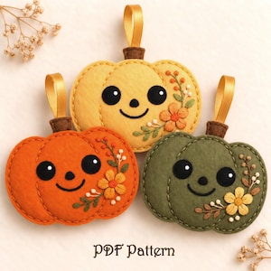 May include: Three felt pumpkin ornaments in yellow, orange, and olive green, each featuring embroidered black eyes, a smiling mouth, and floral accents. Each pumpkin has a brown stem and a gold ribbon loop for hanging. Text reads: PDF Pattern.