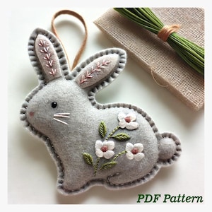 Peut inclure: Une décoration de lapin en feutre gris avec des oreilles brodées roses et des fleurs blanches. Le lapin a un œil noir et des moustaches blanches. La décoration est suspendue à une ficelle brune. Le texte "PDF Pattern" est visible en bas de l'image.