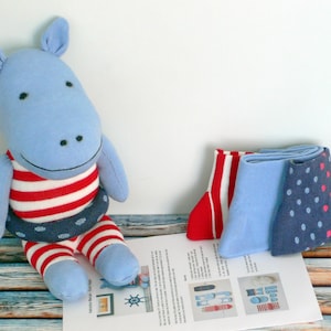 May include: A blue sock hippopotamus wearing a red and white striped swimsuit and a blue and white life preserver. Three socks are shown in the background: one red and white striped sock, one light blue sock, and one navy blue sock with white polka dots.