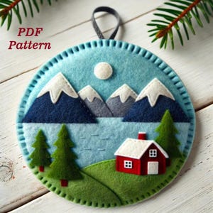 May include: A felt ornament depicting a winter scene. The ornament features a blue lake, mountains, a small red house, and two green trees. The ornament is round and has a white felt moon in the sky.
