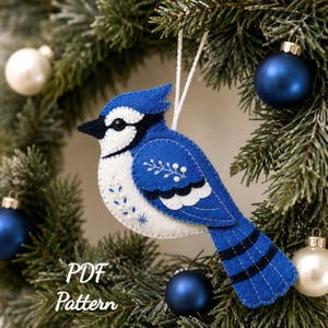 Peut inclure: Une décoration en feutrine représentant un geai bleu avec des coutures blanches et bleues, un bec noir et une ficelle blanche. L'oiseau présente des broderies détaillées sur l'aile et le poitrail. Le texte "PDF Pattern" est visible en bas.