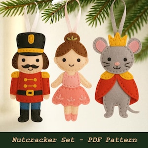 May include: Three felt Christmas ornaments: a nutcracker soldier in red and black, a ballerina in pink, and a gray mouse king with a yellow crown and red cape. The ornaments are hanging from white ribbons.