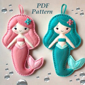 May include: PDF pattern for two felt mermaid ornaments. One mermaid is pink with a pink tail and the other is teal with a teal tail. Both mermaids have a flower on their head and a white top.