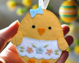 May include: A felt chick ornament with a blue bow, embroidered details, and a white dress with floral embroidery. The chick is yellow with orange feet and a beak.