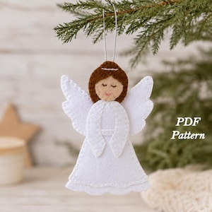 May include: A felt angel ornament with brown hair, white wings, and a white dress, featuring visible stitching along the edges. The figure has closed eyes and a small pink mouth. A silver string is attached to the top. Text reads PDF Pattern.