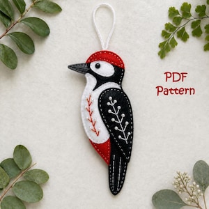 Pode incluir: Um ornamento de feltro em forma de pica-pau com uma crista vermelha, corpo preto e branco e um laço branco para pendurar. O pássaro tem padrões de ramos bordados a branco no peito e na asa, com o texto vermelho PDF Pattern à direita.