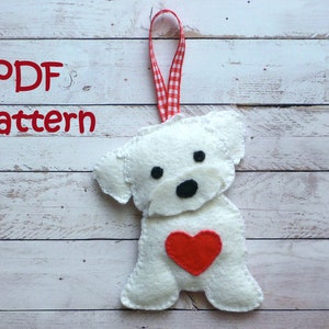 May include: A white felt dog ornament with a red heart on its chest. The dog has black eyes and a black nose. The ornament is hanging from a red and white checkered ribbon. The text "PDF Pattern" is visible in the top left corner of the image.