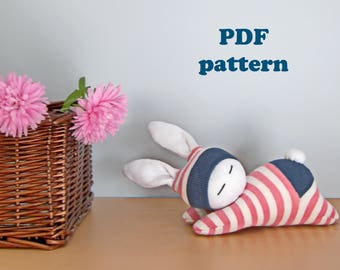 May include: PDF pattern for a sock bunny toy. The bunny is lying on its side with its head resting on its paws. It is made from a red and white striped sock and a blue sock. The bunny has a white face with black eyes and a black mouth.