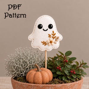 May include: A white felt ghost ornament on a wooden stick featuring black embroidered eyes and a mouth, with a small floral embroidery design in brown and cream. The text PDF Pattern is visible in the upper left corner of the image.