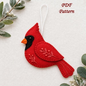Puede incluir: Un adorno de fieltro rojo con forma de cardenal, máscara facial negra, pico naranja y lazo blanco para colgar. El ave está decorada con ramas bordadas y pequeñas cuentas rojas en el ala y el cuerpo. El texto PDF Pattern es visible.