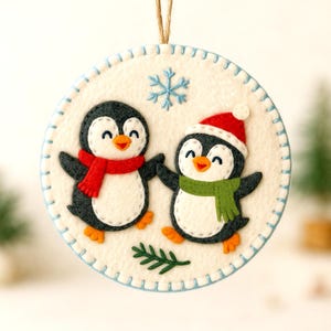 Op de afbeelding: Een ronde, witte vilten kerstversiering met twee lachende pinguïnfiguren. Eén pinguïn draagt een rode sjaal, de andere een groene sjaal en een rode muts. Een blauwe sneeuwvlok en een takje groen zijn ook aanwezig.