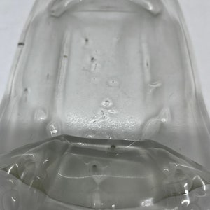 May include: A clear, transparent glass object with a rounded top and a flared base. The glass has a slightly textured surface with small imperfections. The object appears to be a container or a part of a larger vessel.