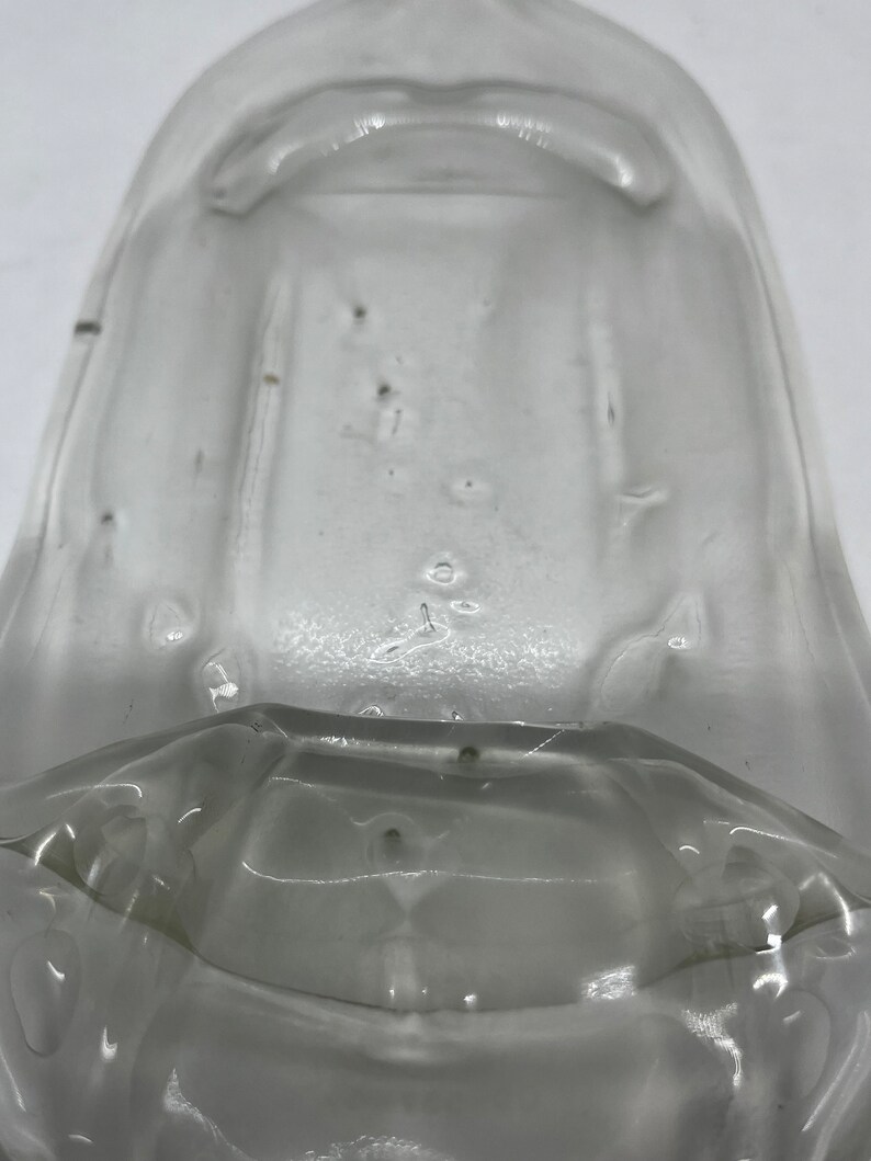 May include: A clear, transparent glass object with a rounded top and a flared base. The glass has a slightly textured surface with small imperfections. The object appears to be a container or a part of a larger vessel.
