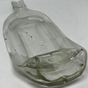 May include: A clear, antique glass bottle with a flattened, rectangular body and a narrow neck. The bottle has a slightly curved shape and appears to be hand-blown, with visible imperfections in the glass. The bottle is empty and sits on a white surface.