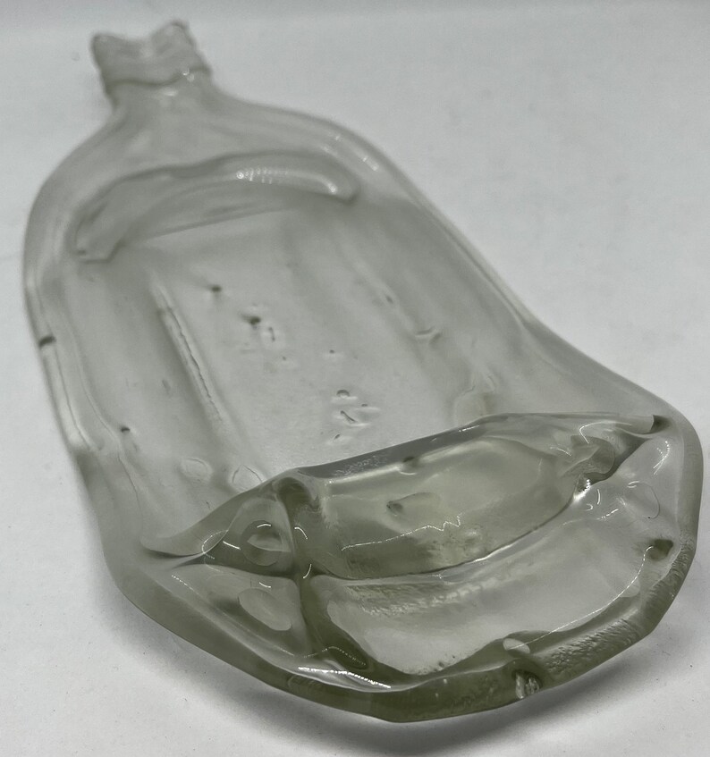 May include: A clear, antique glass bottle with a flattened, rectangular body and a narrow neck. The bottle has a slightly curved shape and appears to be hand-blown, with visible imperfections in the glass. The bottle is empty and sits on a white surface.