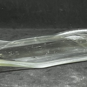 May include: A clear, flattened glass bottle, reshaped into a serving dish. The bottle's original form is still visible, with the neck and base intact. The glass has a slight green tint, and the dish is resting on a dark surface.