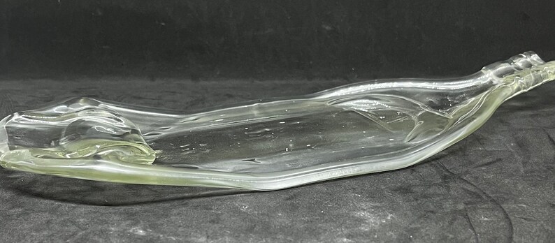 May include: A clear, flattened glass bottle, reshaped into a serving dish. The bottle's original form is still visible, with the neck and base intact. The glass has a slight green tint, and the dish is resting on a dark surface.