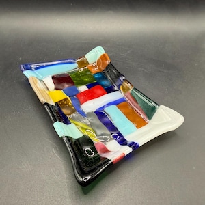 May include: A rectangular, fused glass dish with a woven pattern of colorful glass strips. The dish features a mix of colors including blue, red, yellow, green, and white. The edges are slightly curved, and the dish is on a dark background.