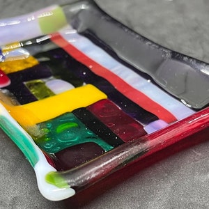 Pode incluir: Um pequeno prato retangular de vidro com um design abstrato colorido. O prato é feito de vidro fundido e tem uma variedade de cores, incluindo vermelho, amarelo, verde, azul e roxo.