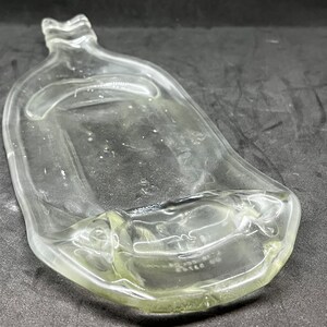 May include: A clear, glass bottle with a unique, curved shape. The bottle has a wide, flat body and a narrow neck with a decorative top. The glass appears to be slightly tinted, and the bottle is empty.