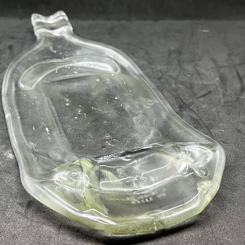 May include: A clear, glass bottle with a unique, curved shape. The bottle has a wide, flat body and a narrow neck with a decorative top. The glass appears to be slightly tinted, and the bottle is empty.