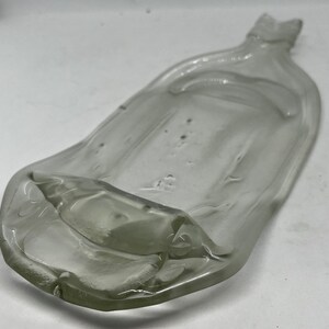 May include: A clear, glass, shallow dish with a unique, elongated shape. The dish has a slightly raised edge and a small, pointed extension at one end. The glass has a few small bubbles and imperfections.