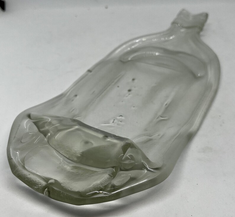 May include: A clear, glass, shallow dish with a unique, elongated shape. The dish has a slightly raised edge and a small, pointed extension at one end. The glass has a few small bubbles and imperfections.