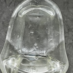 May include: Clear glass flask with a wide, flat body and a narrow neck. The flask has a rectangular base and a slightly curved top. The glass has some imperfections and bubbles. The flask is empty and appears to be vintage.