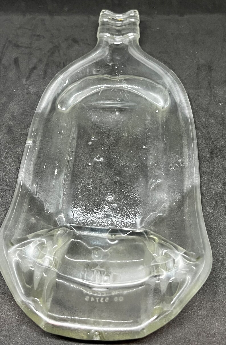 May include: Clear glass flask with a wide, flat body and a narrow neck. The flask has a rectangular base and a slightly curved top. The glass has some imperfections and bubbles. The flask is empty and appears to be vintage.
