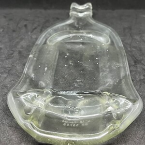 May include: A clear, bell-shaped glass bottle with a flared base and a decorative top. The bottle has a slightly yellow tint and appears to be empty. The base of the bottle has some text. The overall design is simple and elegant.