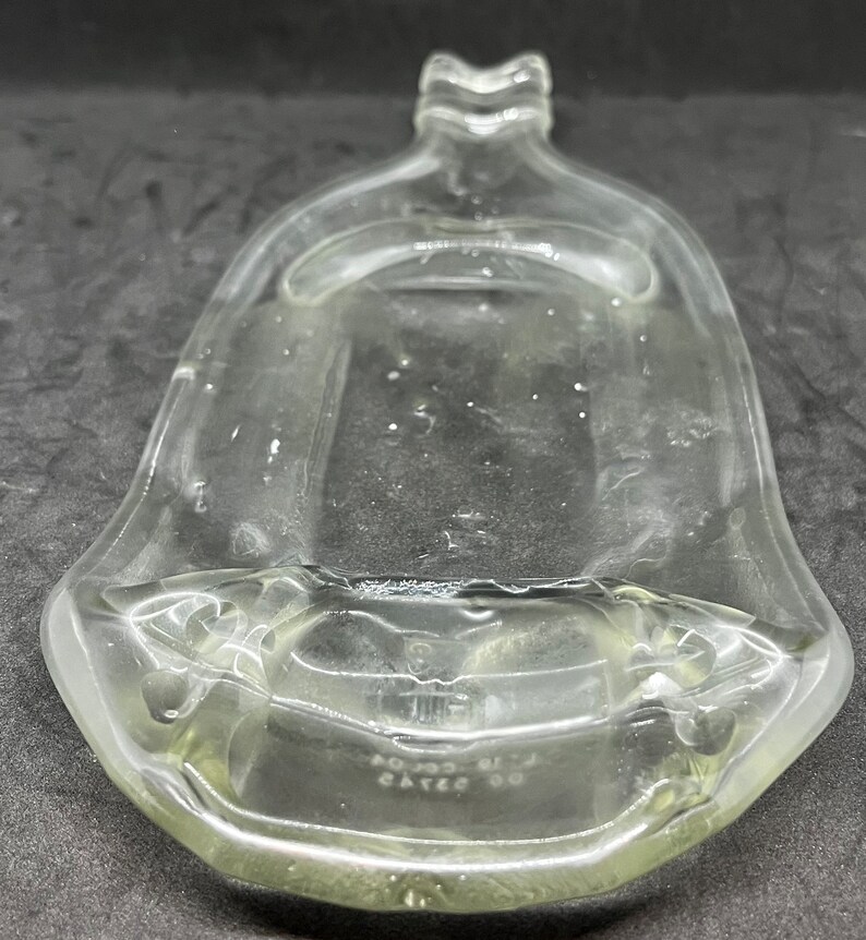 May include: A clear, bell-shaped glass bottle with a flared base and a decorative top. The bottle has a slightly yellow tint and appears to be empty. The base of the bottle has some text. The overall design is simple and elegant.
