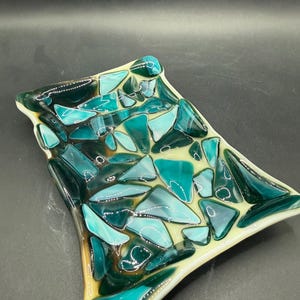 Könnte beinhalten: Eine dekorative Glasschale mit Mosaikdesign. Die Schale zeigt eine Mischung aus türkis, türkis und dunkelgrünen Glasstücken auf einem hellgelben Untergrund. Die Ränder sind hellgelb umrandet.