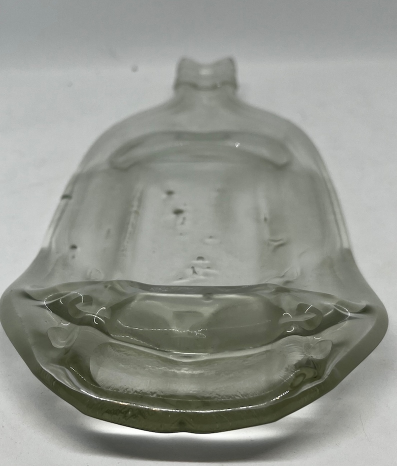 May include: A clear, antique glass bottle with a wide, rounded base and a narrow neck. The bottle has a unique, curved shape and appears to be made of thick glass. The bottle's design suggests it may have been used for liquids.