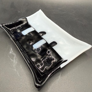 May include: A rectangular glass dish with a divided design. One side is glossy black with white accents, and the other is white. The dish has a slightly curved shape and is likely used for decorative purposes or as a small serving tray.