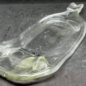 May include: A clear, glass bottle with a unique, elongated shape. The bottle has a wide, flat base that tapers to a narrow neck and a handle-like top. The glass has a slight greenish tint and appears to be vintage.