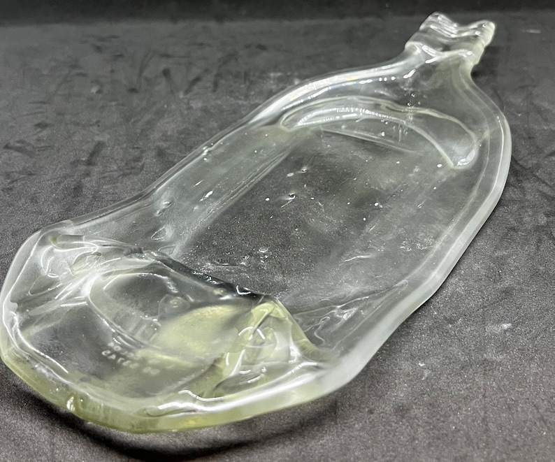 May include: A clear, glass bottle with a unique, elongated shape. The bottle has a wide, flat base that tapers to a narrow neck and a handle-like top. The glass has a slight greenish tint and appears to be vintage.