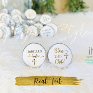 Puede incluir: Dos etiquetas redondas blancas con letras doradas. Una etiqueta dice "ISABELLE'S dedication" con una cruz dorada. La otra etiqueta dice "Bless THIS Child" con una cruz dorada. Las etiquetas están sobre una superficie blanca y esponjosa.