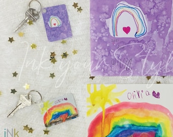 Könnte beinhalten: Zwei Schlüsselanhänger mit Kinderkunstwerken von Regenbögen. Der erste Schlüsselanhänger hat einen lila Hintergrund mit einem Regenbogen und einem Herzen. Der zweite Schlüsselanhänger hat einen Regenbogen mit einer Sonne und einem Herzen. Beide Schlüsselanhänger tragen den Namen "Olivia".
