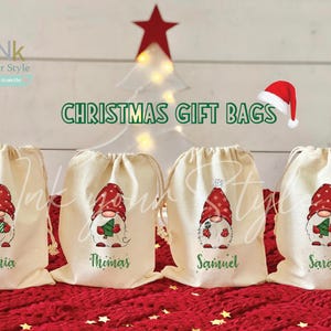 May include: Four white Christmas gift bags with drawstring closures. Each bag features a different gnome design with a red hat, white beard, and a Christmas tree. The bags are labeled with the names Sophia, Thomas, Samuel, and Sarah.
