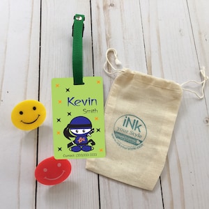 May include: Green luggage tag with a cartoon ninja, the name Kevin Smith, and a phone number. The tag is on a light wood surface with a yellow smiley face and a red smiley face. There is a small white drawstring bag with a green logo that says "ink your style" and a website address.