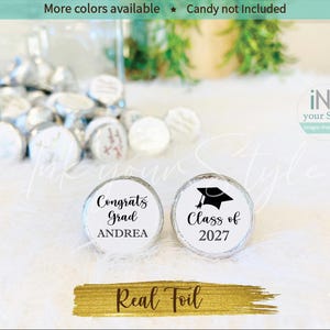 May include: Two round silver foil candy stickers featuring graduation themes. One sticker displays the text Congrats Grad ANDREA, while the other shows a black graduation cap icon with the text Class of 2027. The stickers are shown in front of a glass jar.