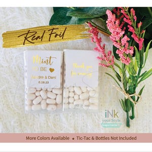 May include: Two small white boxes filled with white Tic Tacs. The boxes have gold foil labels. One label says "Mint to be" with a heart and the names "andra & Dani" and the date "8.28.23". The other label says "Thank you for coming".