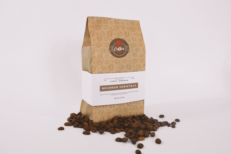 May include: A brown paper bag with a coffee bean pattern and a white label that reads "HAND CRAFTED COPAN, HONDURAS BOURBON VARIETALS NET WT 12 OZ". The bag is sitting on a white surface with coffee beans scattered around it.