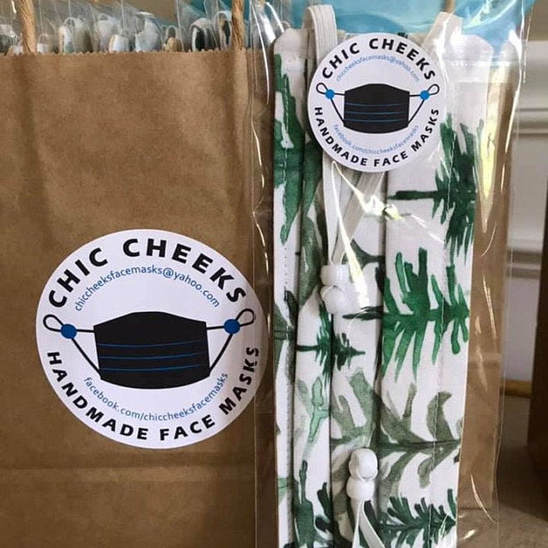 May include: A white fabric face mask with a green leafy pattern. The mask is packaged in a clear plastic bag with a white elastic band. The bag has a sticker that says "Chic Cheeks Handmade Face Masks" with a black and blue face mask illustration. The sticker also includes the website address "chiccheekfacemasks@yahoo.com" and the social media handle "facebook.com/chiccheekfacemasks".