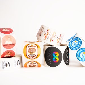 May include: Rolls of round stickers with various designs.  One roll features a donut with the text "Donuts Creamy Est'd 1995". Another roll features a rainbow infinity symbol with the text "Infinite Creative Design". A third roll features a blue wave with the text "Surfing Club Beckly Beach".